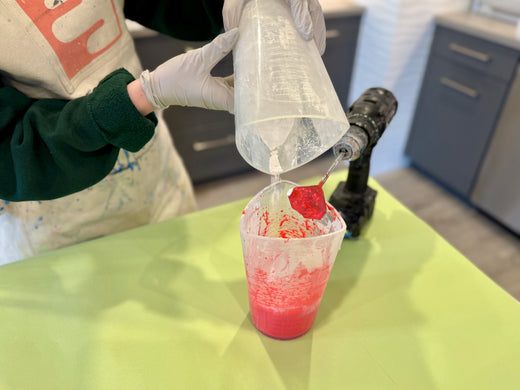 Person wearing gloves and an apron pouring a Jesmonite AC100 powder base into a clear container with red-colored Jesmonite AC100 liquid, preparing a mixture on a green workspace. A drill with a high-sheer mixing blade is visible in the background.